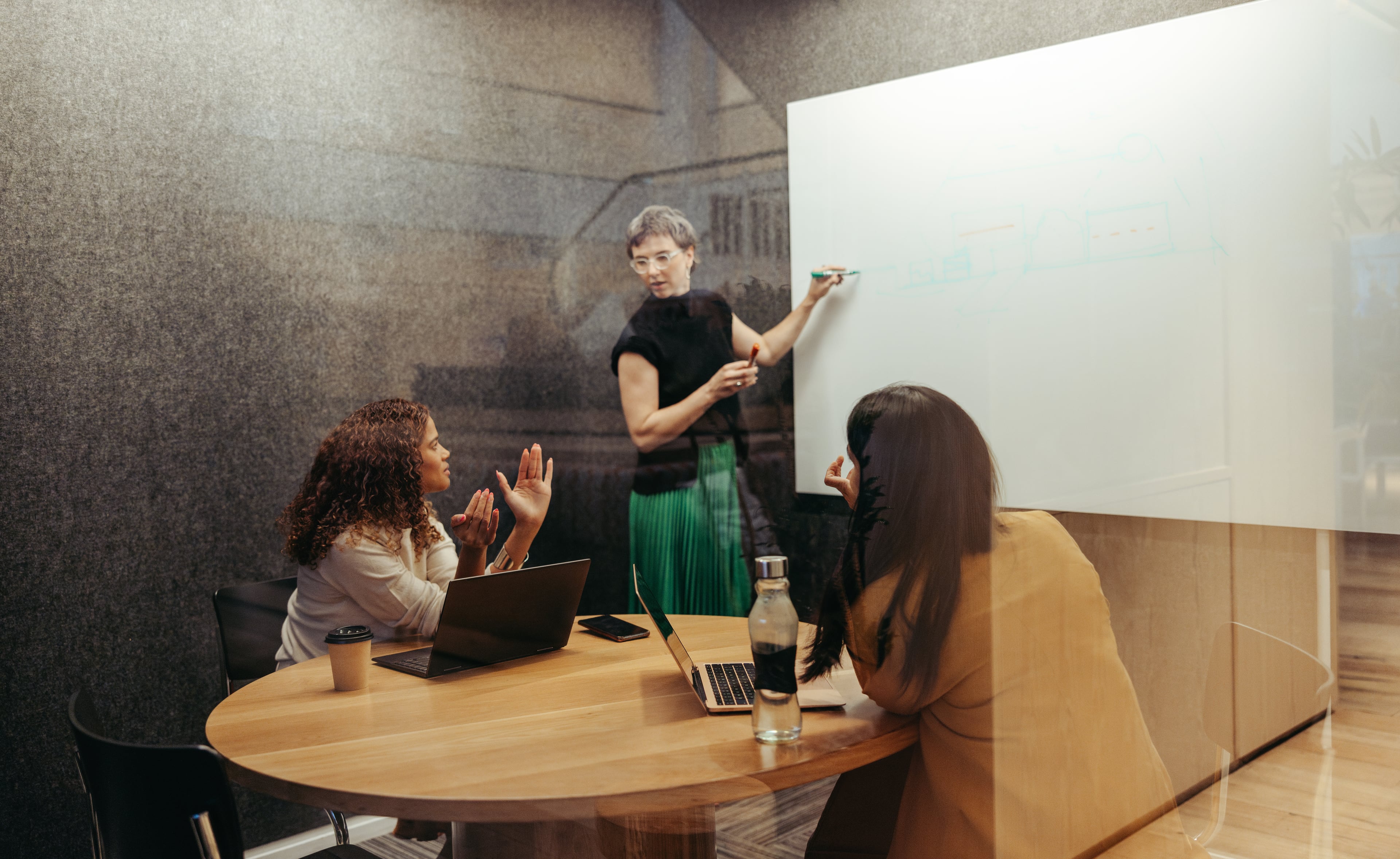 Drei Personen in einem Meetingraum, zwei sitzen am Tisch mit Laptops, eine steht am Whiteboard und erklärt etwas.