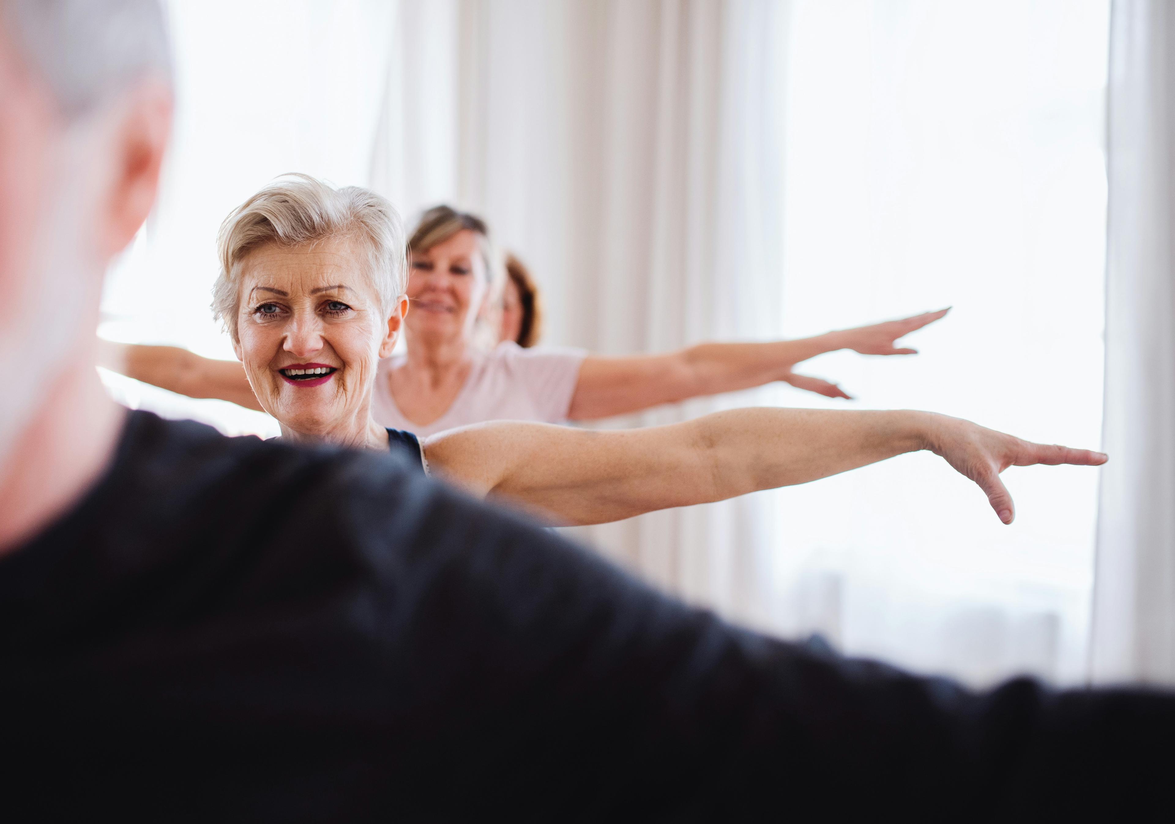 Living Longer – but with Quality Older adults stretching together in a bright room. A smiling woman in the foreground captures a moment of movement, connection, and joy in later life.