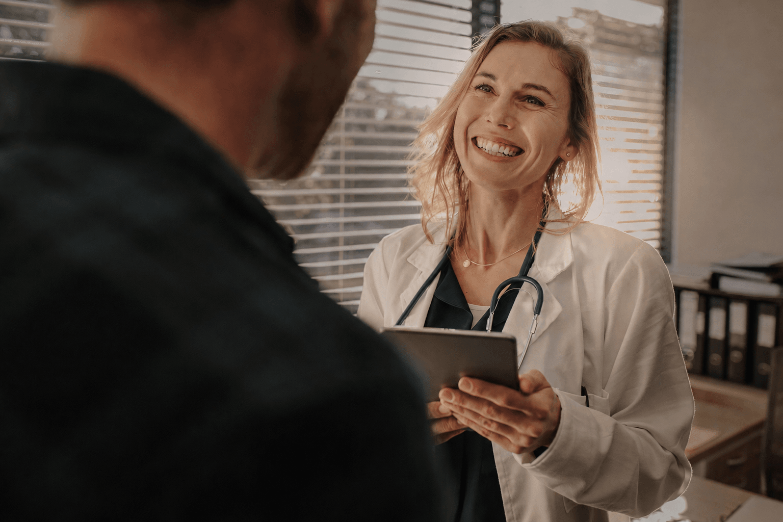 Smiling doctor holding a tablet while speaking with a patient during an in-person consultation in a medical office.
