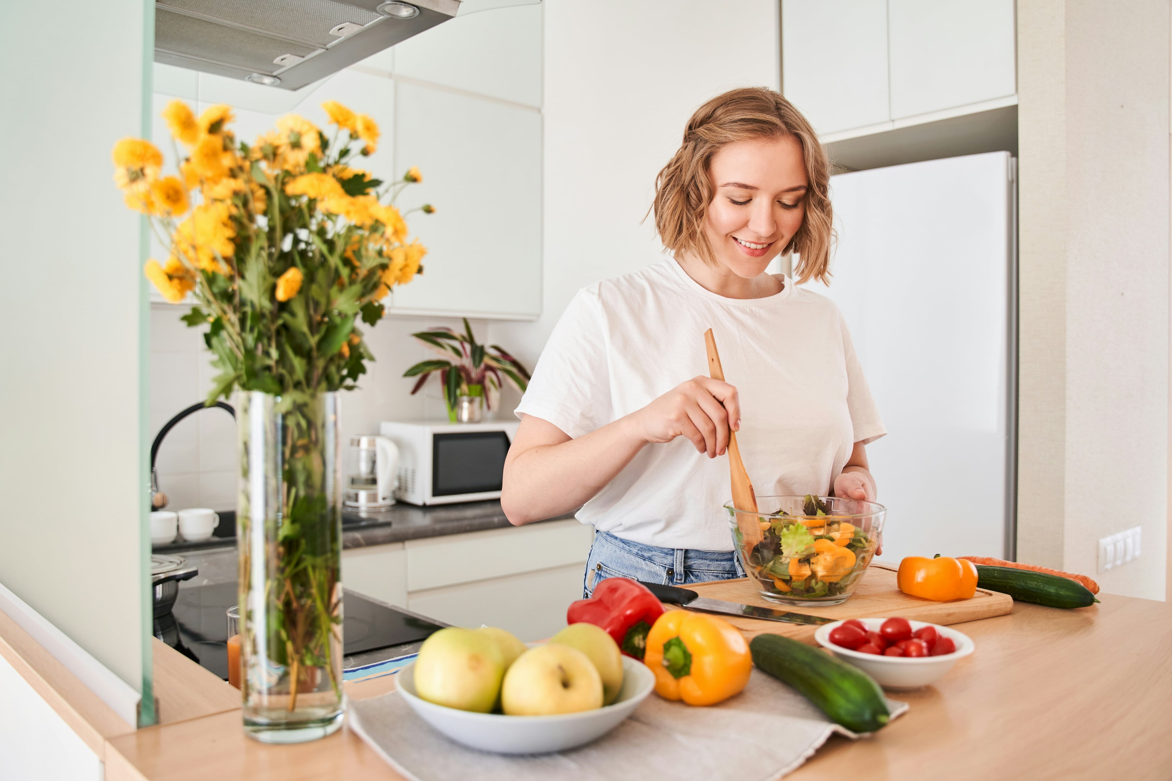Junge Frau bereitet in einer hellen Küche einen bunten Salat zu. Auf der Küchenzeile liegen frisches Obst und Gemüse, im Vordergrund steht eine Vase mit gelben Blumen.