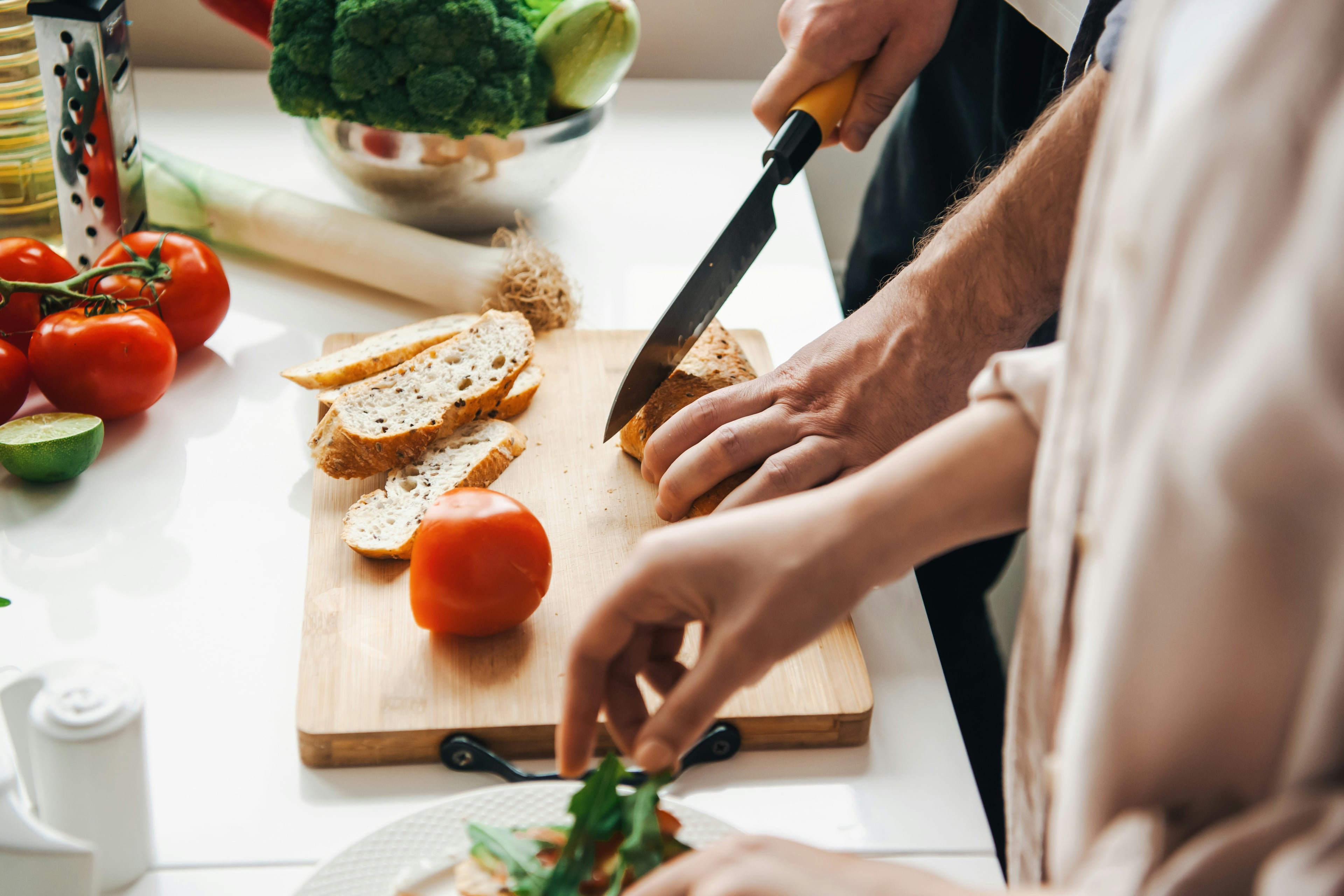 Zwei Personen bereiten gemeinsam eine Mahlzeit in der Küche zu. Eine Hand schneidet Brot auf einem Holzbrett, während die andere frisches Gemüse garniert. Umgeben von Tomaten, Lauch und Brokkoli.