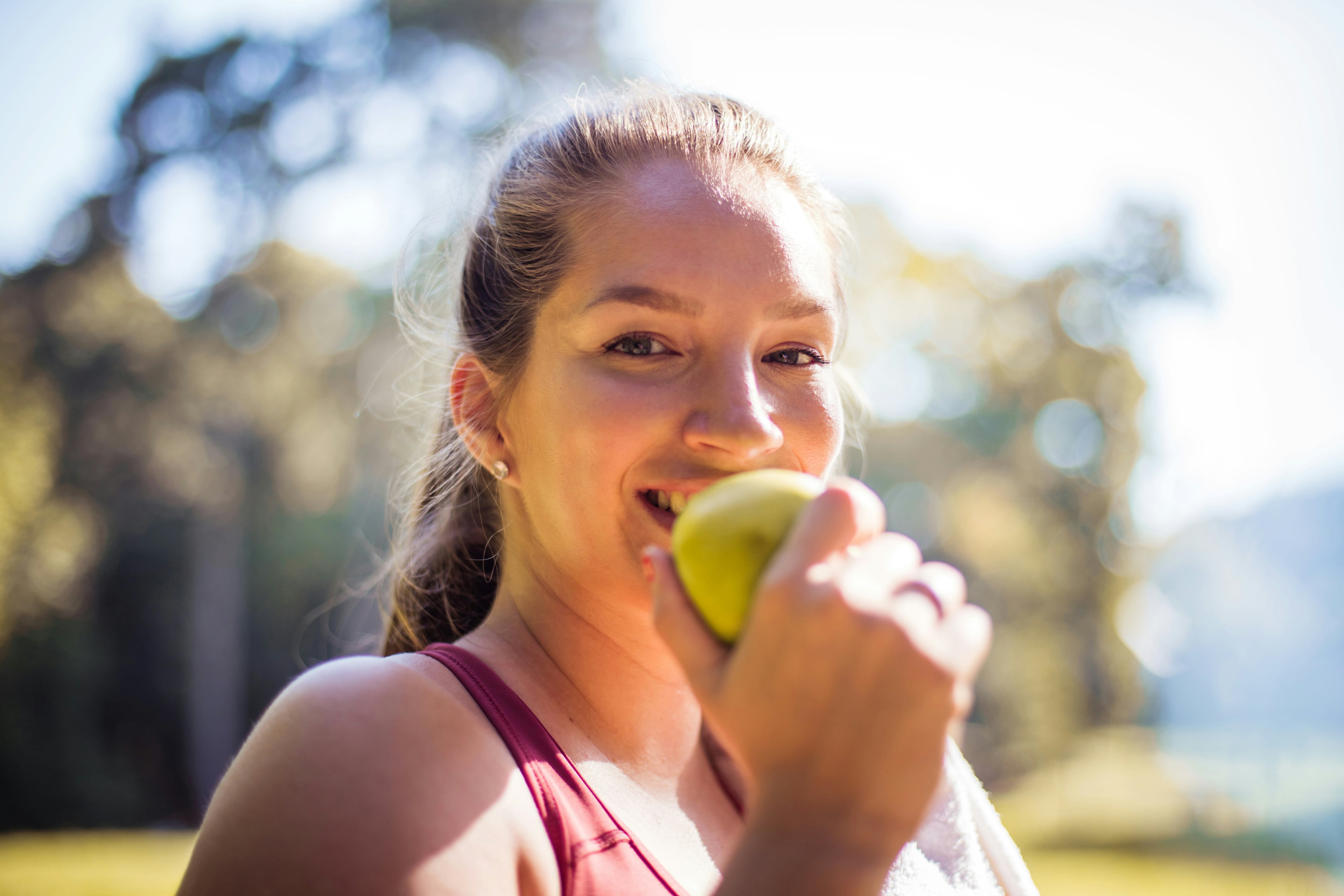 Junge Frau beißt lächelnd in einen grünen Apfel. Sie trägt Sportkleidung und hat ein Handtuch über der Schulter – Szene im Freien bei Sonnenschein.