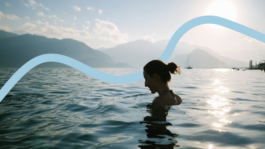 A woman swims in a calm mountain lake at sunset, with mountains and sailboats visible in the background.