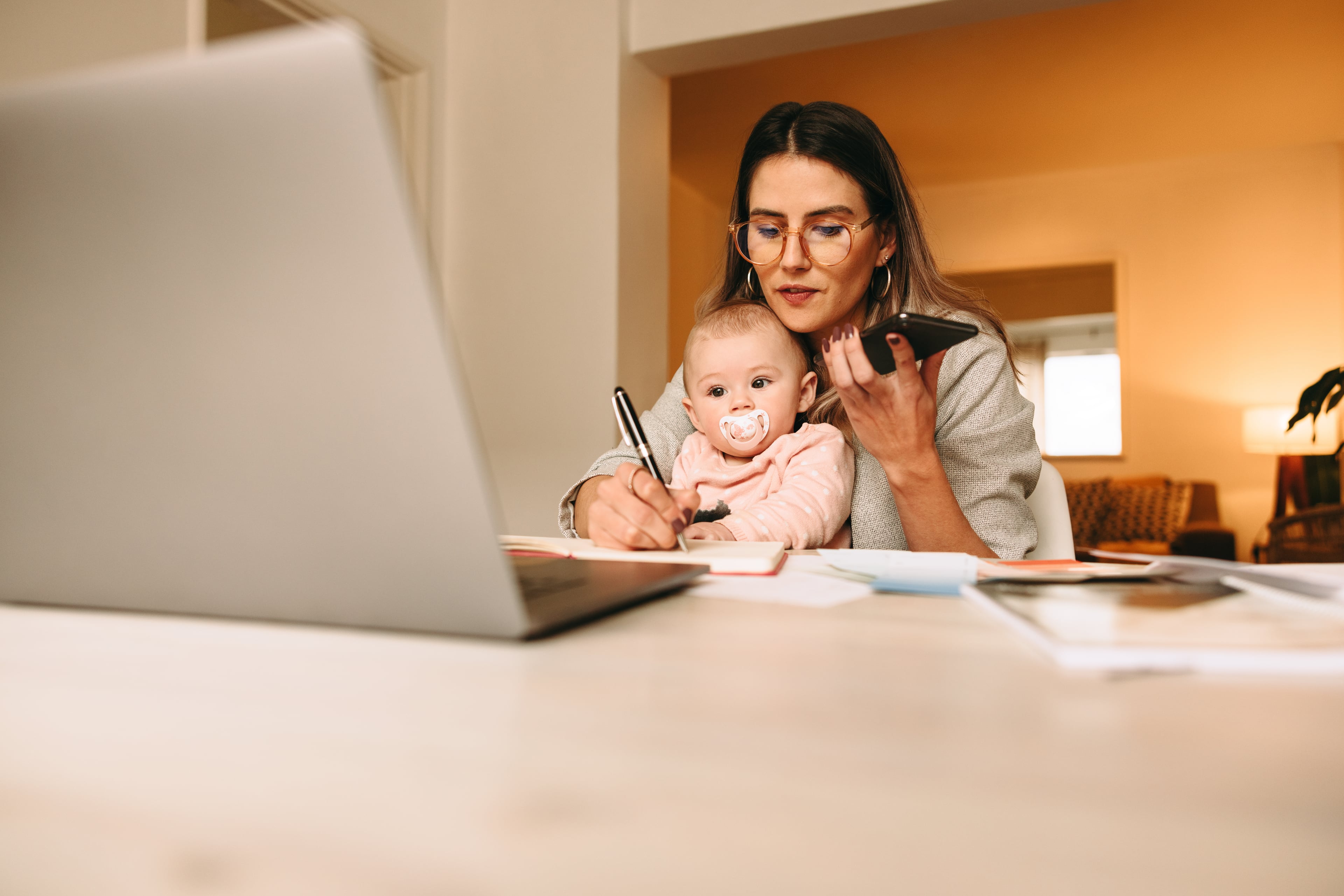 Mother working from home with a baby while using a laptop and smartphone.