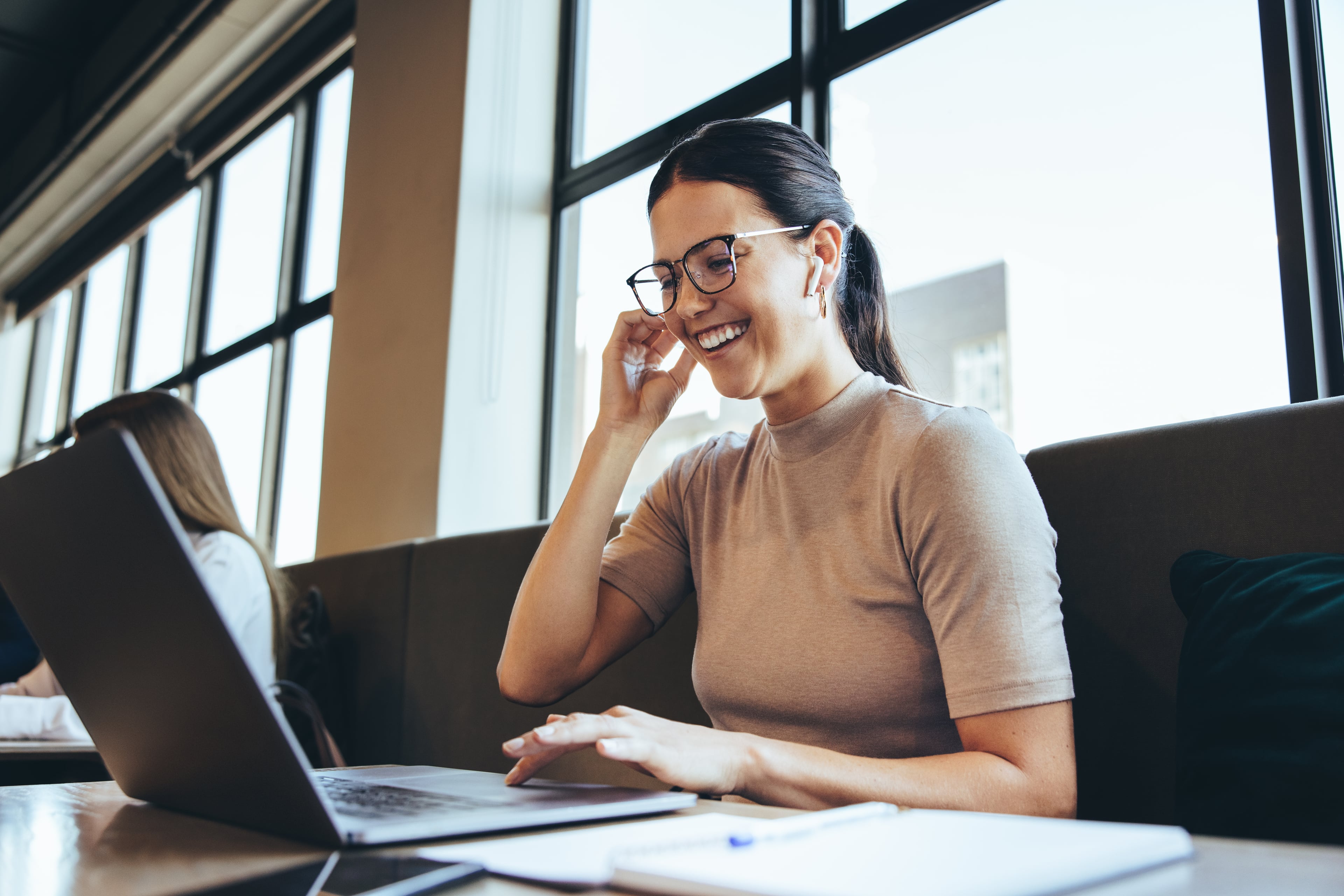 Eine junge Frau mit Brille sitzt lächelnd in einem modernen Café oder Co-Working-Space vor ihrem Laptop und telefoniert über Kopfhörer.