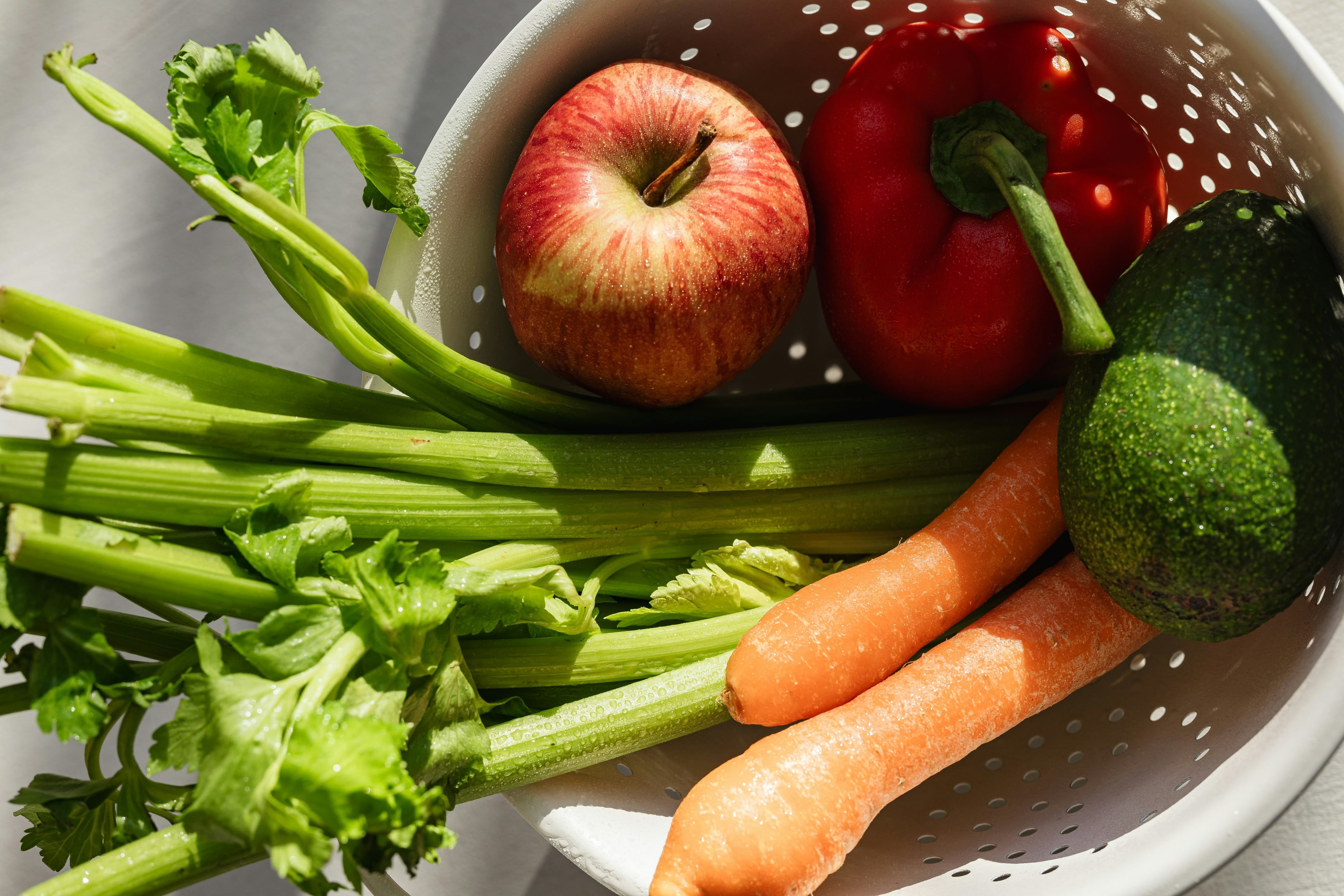 Nahaufnahme eines weißen Siebs mit frischem Gemüse und Obst: Stangensellerie, Karotten, Apfel, Paprika und Avocado – symbolisiert gesunde Ernährung.