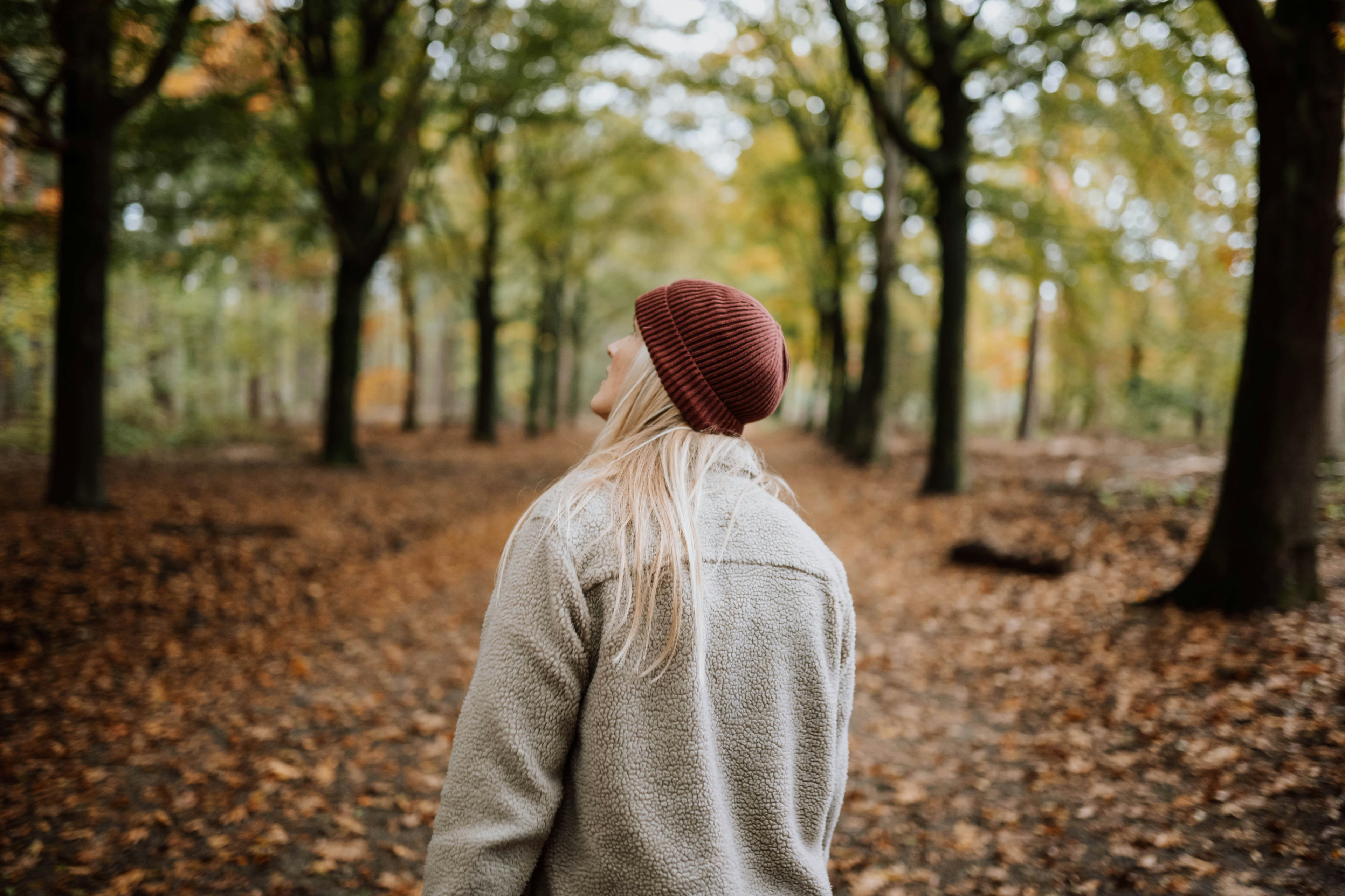 Woman in a rust-red beanie and sherpa fleece jacket looks up on a leaf-covered forest path in autumn, photographed from behind
