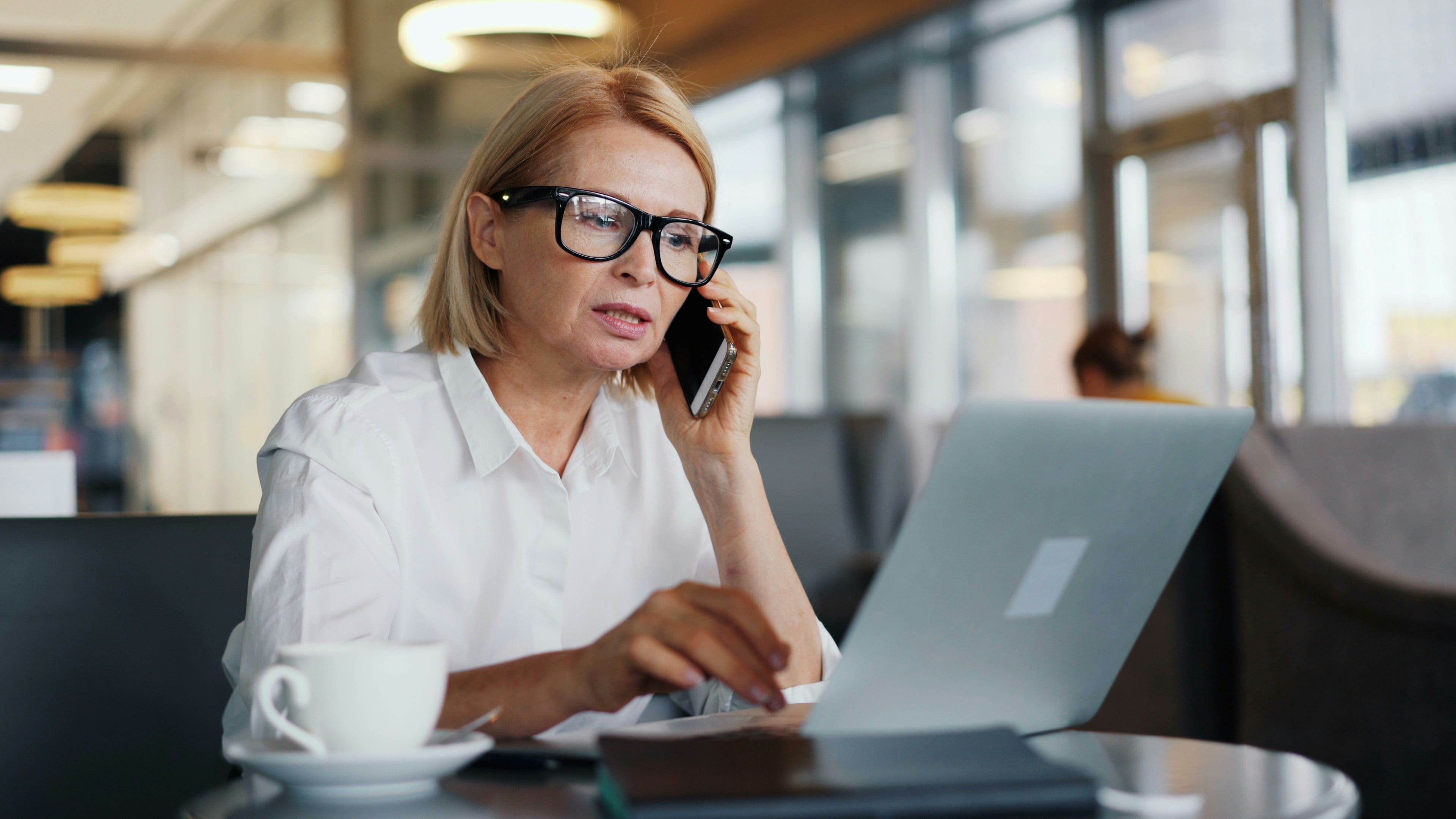 A woman with a blonde bob and black glasses sits at a laptop in a café or lobby, talking on her smartphone while typing at the same time, with a coffee cup on the table beside her.