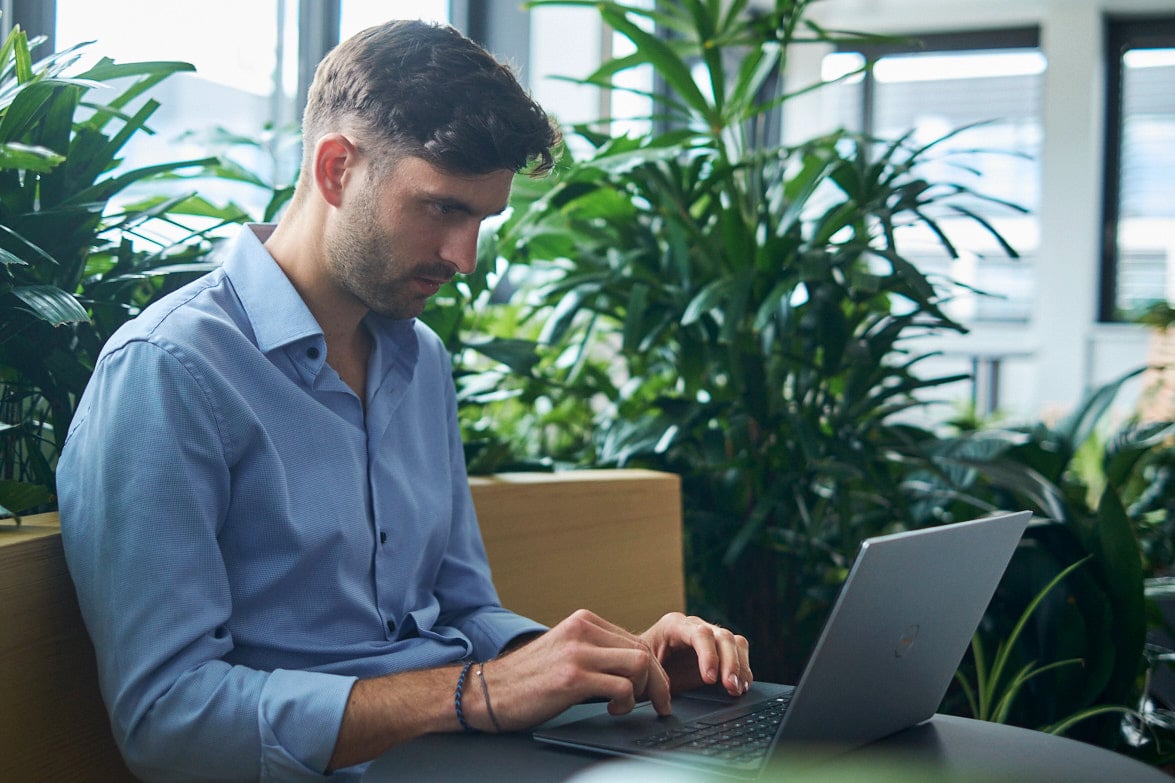 Man working on a laptop in a modern office surrounded by green plants.