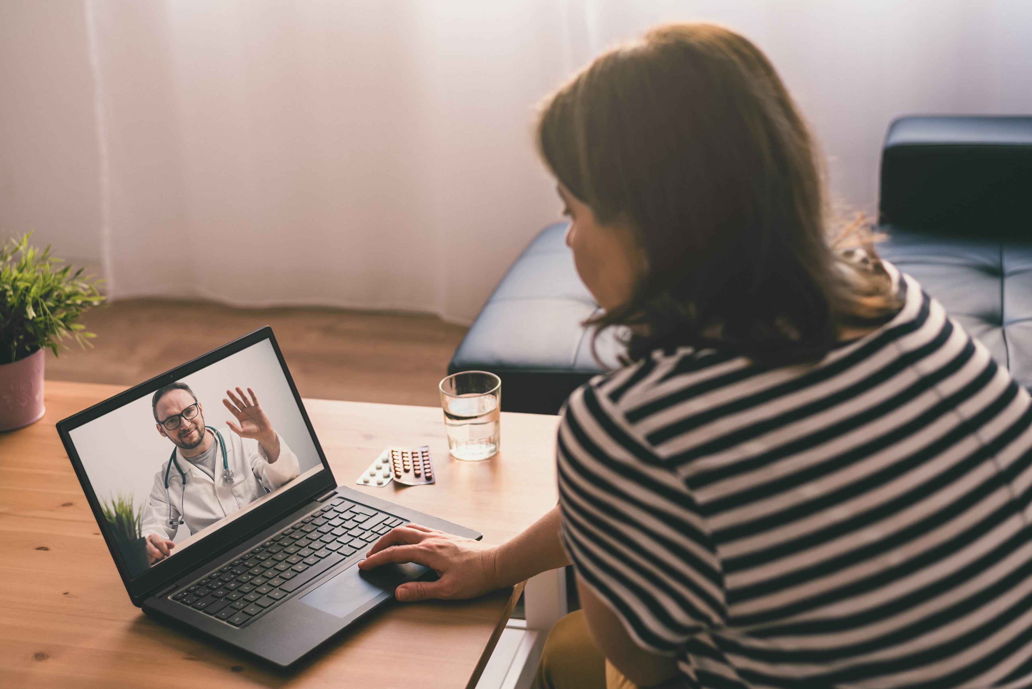 A woman in a striped shirt is having a video consultation with a doctor on her laptop in a living room.