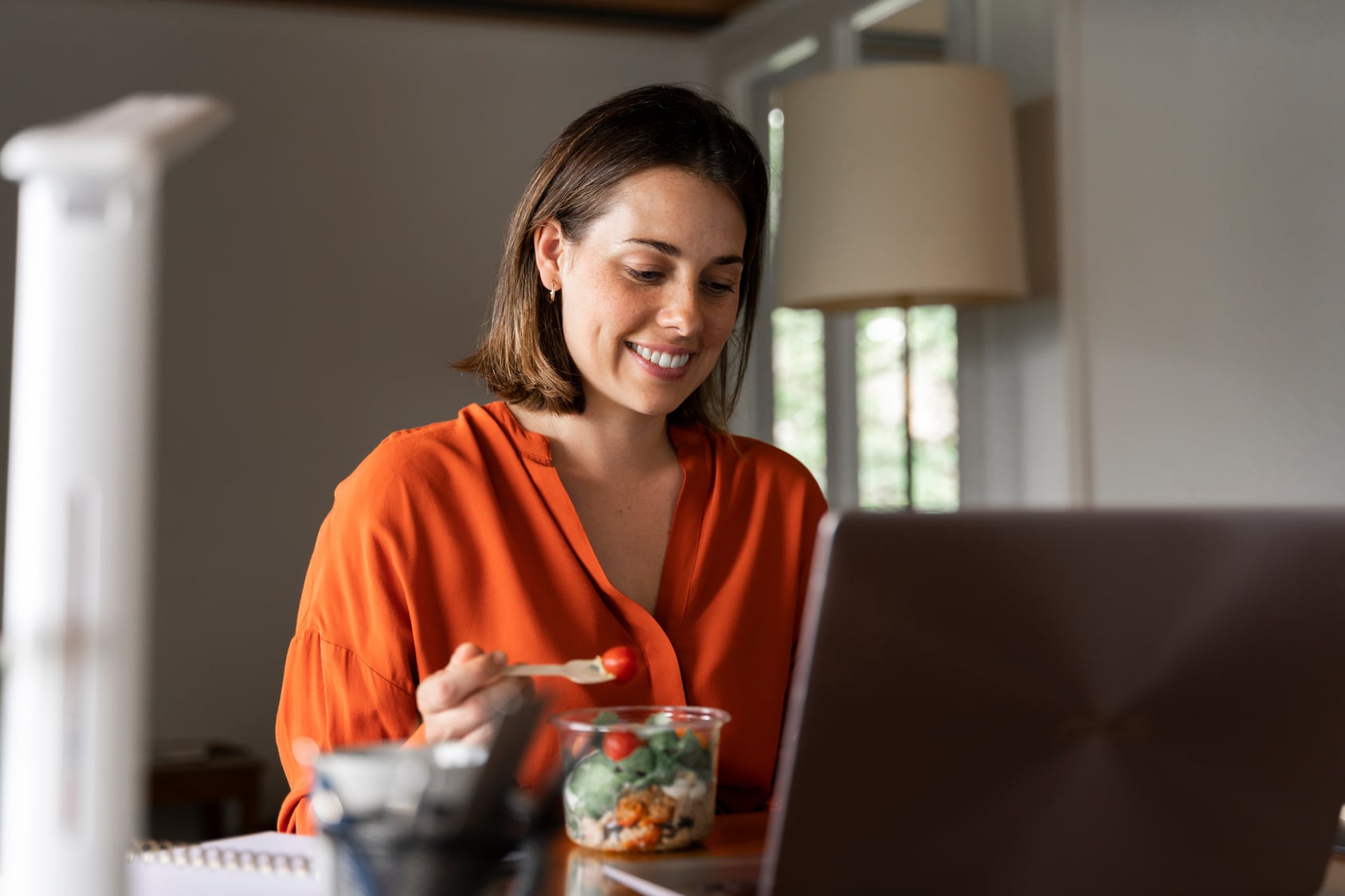 Ernährung 4 Frau in orangefarbener Bluse sitzt lächelnd vor einem Laptop und isst dabei einen frischen Salat aus einer Schale – eine entspannte Arbeitspause im Homeoffice oder Büro.