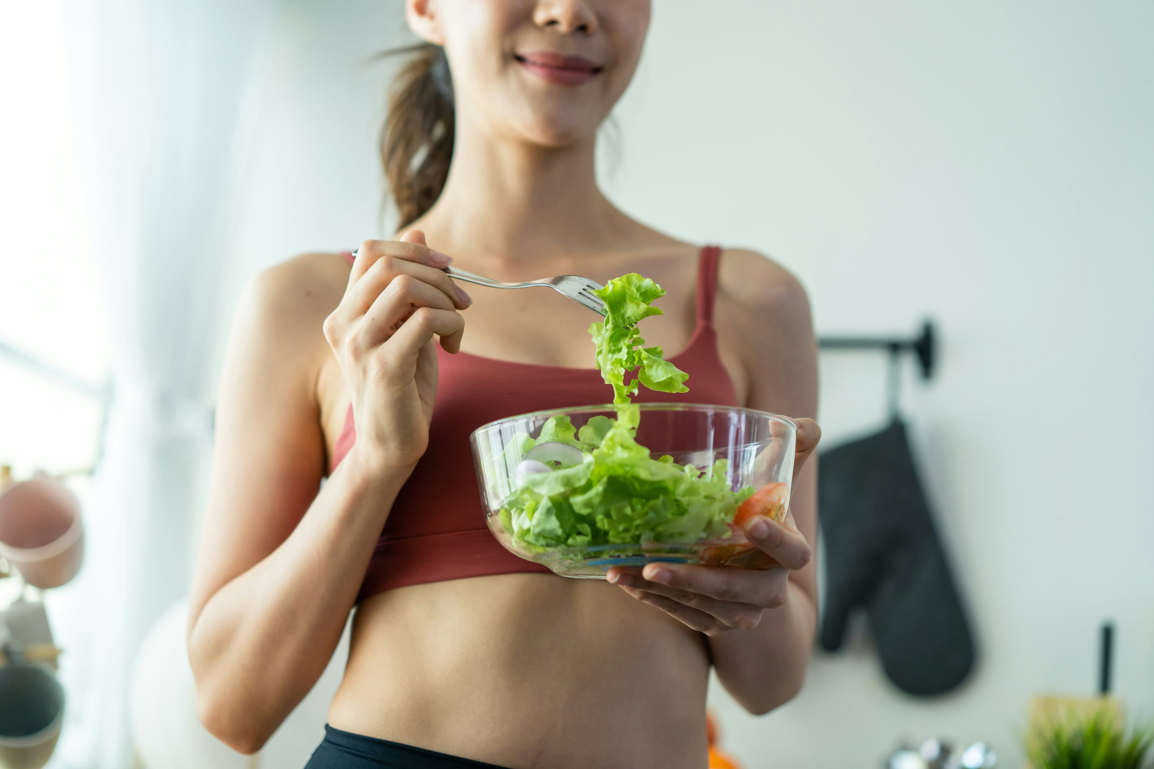 Junge Frau in Sportkleidung hält eine Glasschüssel mit frischem Salat und isst mit einer Gabel – gesunde Ernährung in entspannter Atmosphäre.