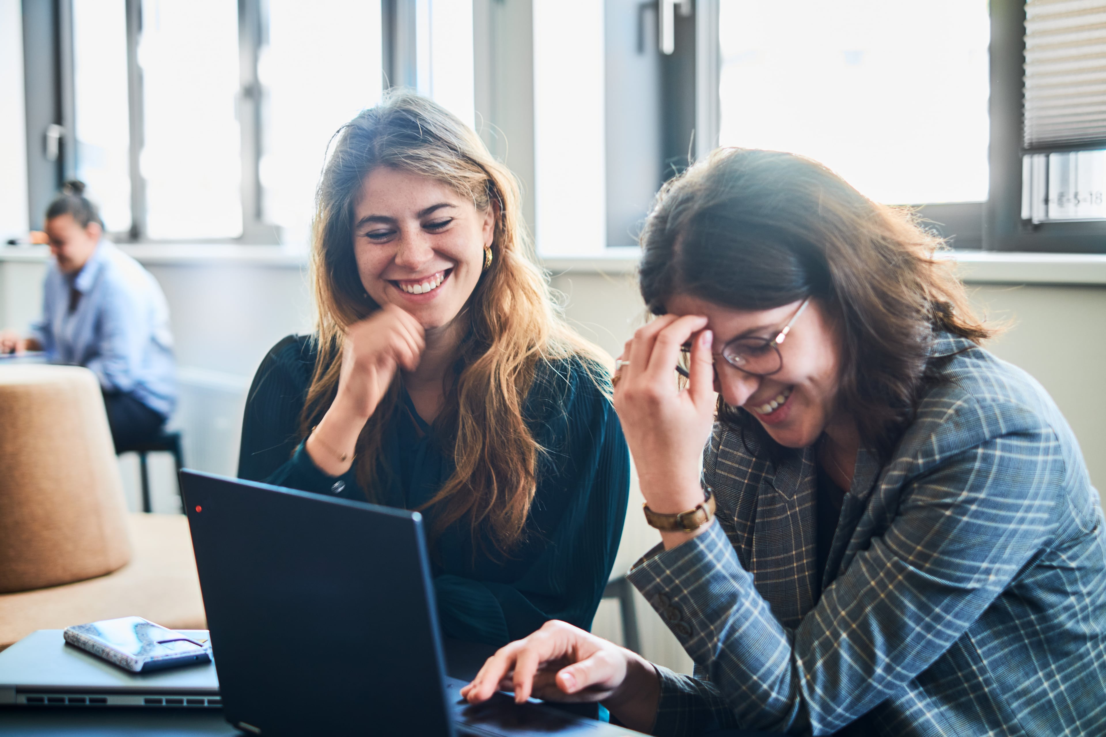 Zwei junge Frauen sitzen gemeinsam vor einem Laptop und lachen herzlich – eine entspannte und kollegiale Arbeitssituation in einem modernen Büro.