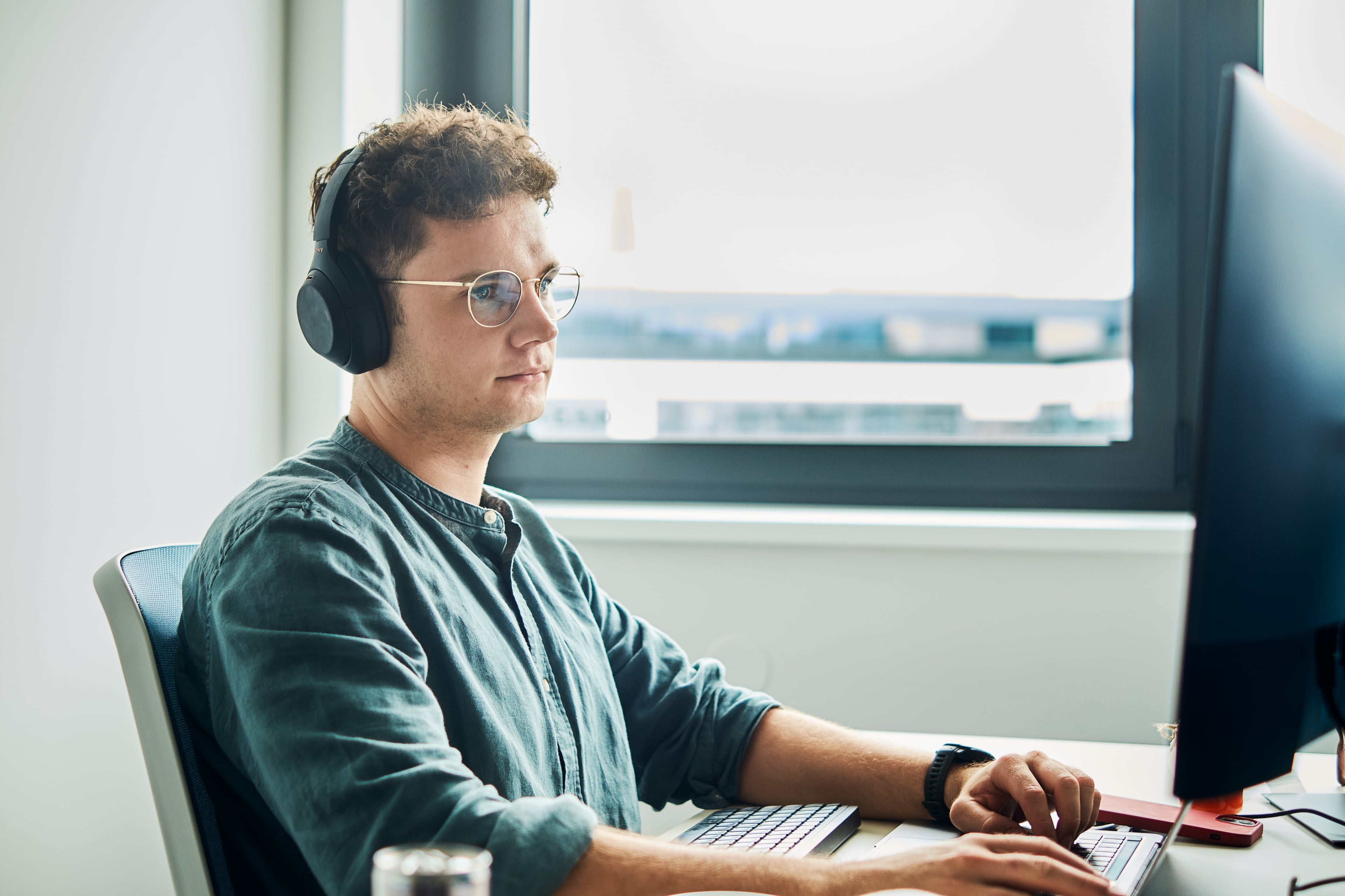 Mann mit Kopfhörern und Brille sitzt an einem Schreibtisch und arbeitet konzentriert am Computer. Durch das Fenster fällt Tageslicht in den modernen Büroraum.