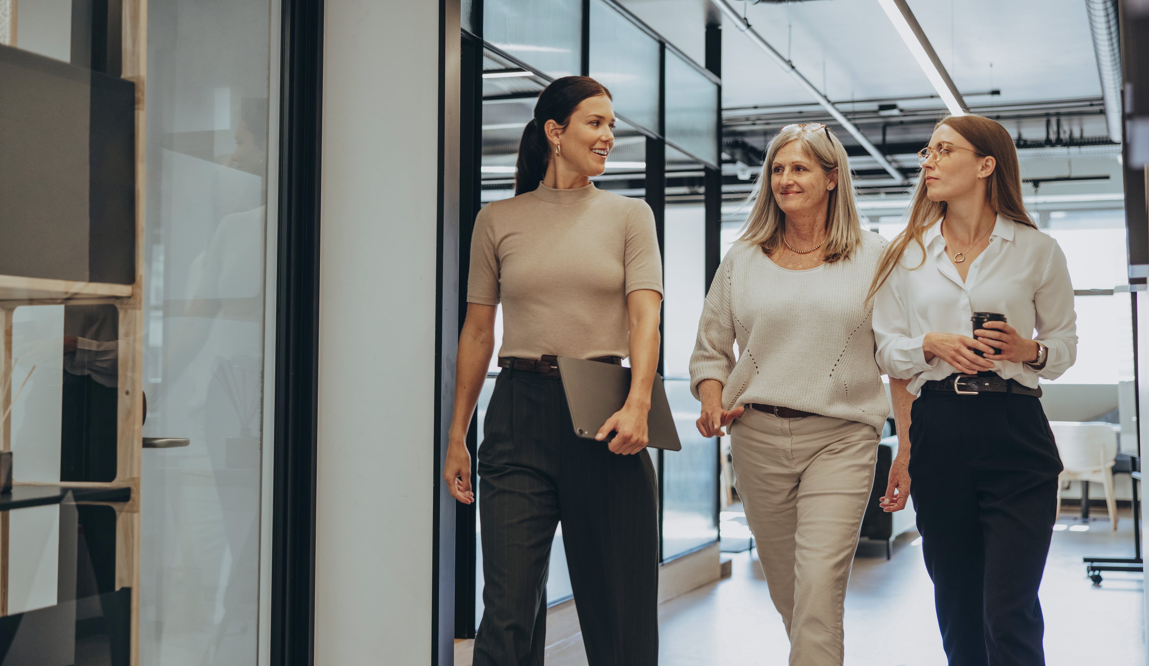 Drei Frauen gehen gemeinsam durch ein modernes Büro und unterhalten sich freundlich. Eine trägt ein Tablet, eine andere ein Smartphone – die Szene wirkt professionell und dynamisch.