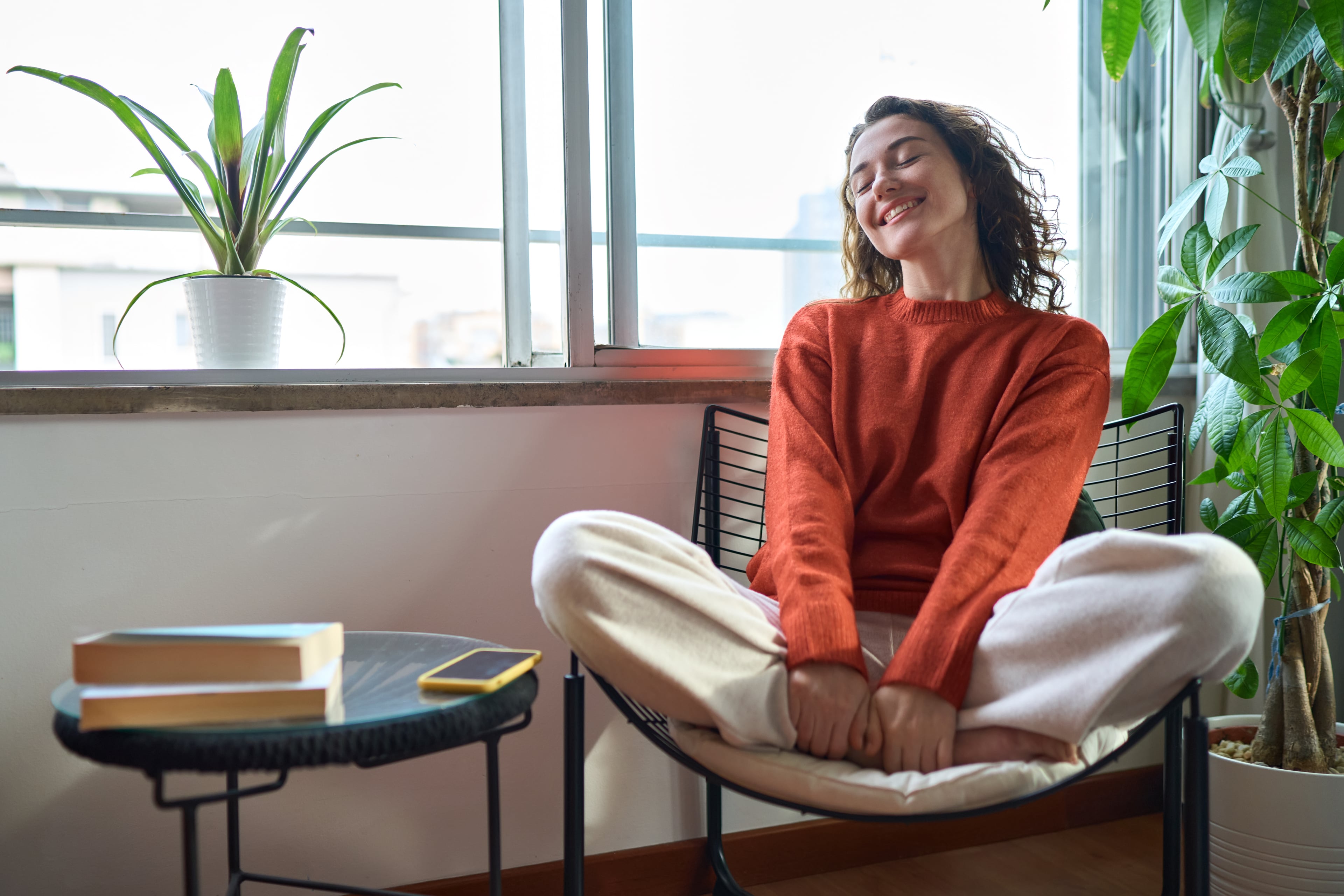 A woman sitting comfortably on a chair by a window, smiling with closed eyes and enjoying a calm moment in a bright space.