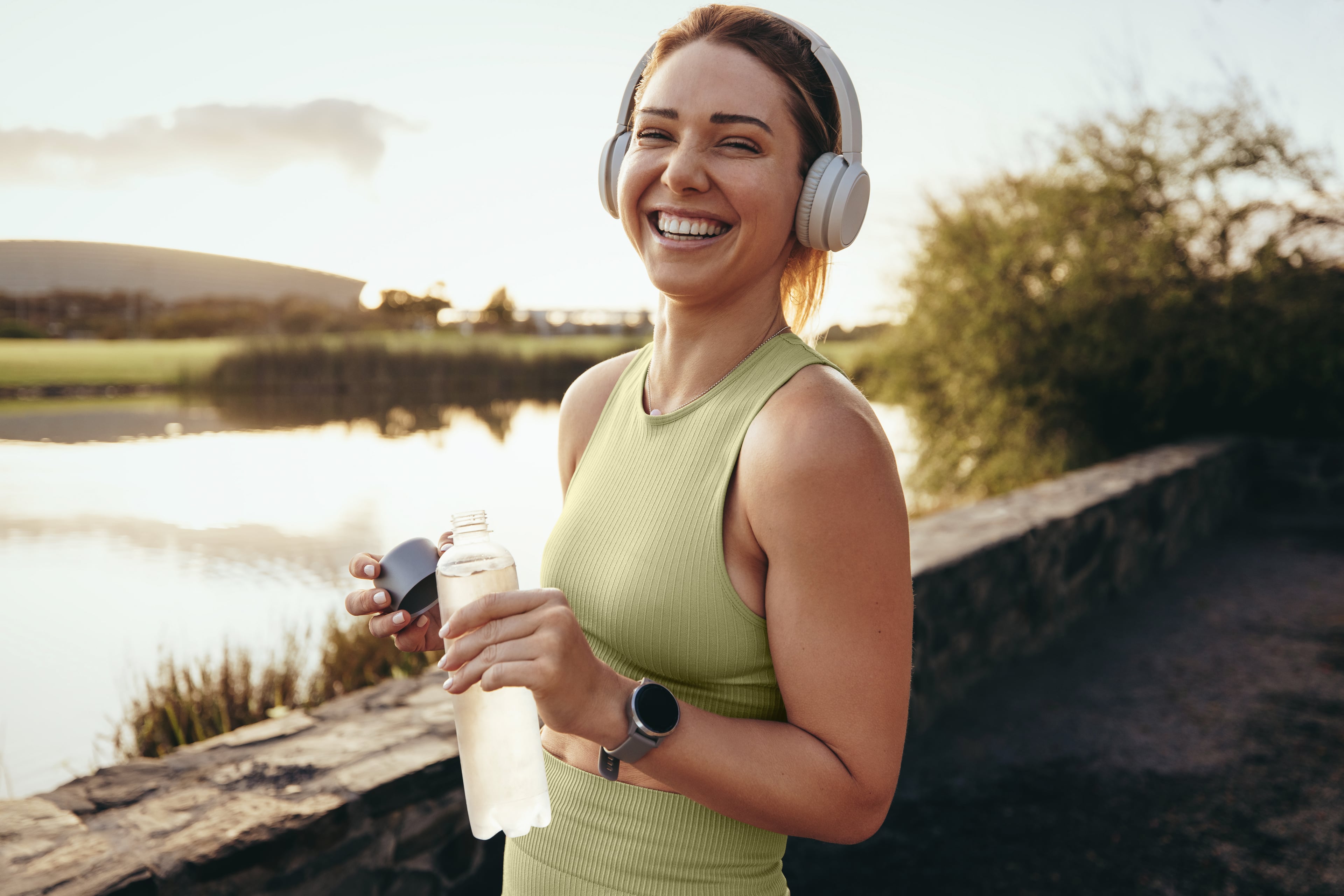 Eine Person steht vor einer Naturlandschaft und trägt sportliche Kleidung. Sie hält eine Wasserflasche in der Hand.