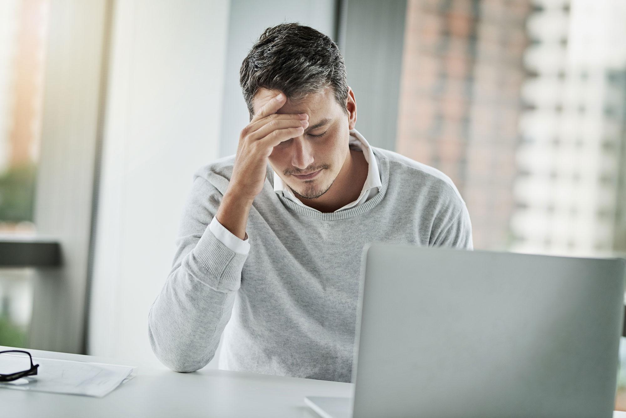 AdobeStock 488093166 gestresster-mann Man sitting in front of a laptop in a modern office, holding his forehead thoughtfully. A pair of glasses and documents lie next to him – he appears stressed or overwhelmed.