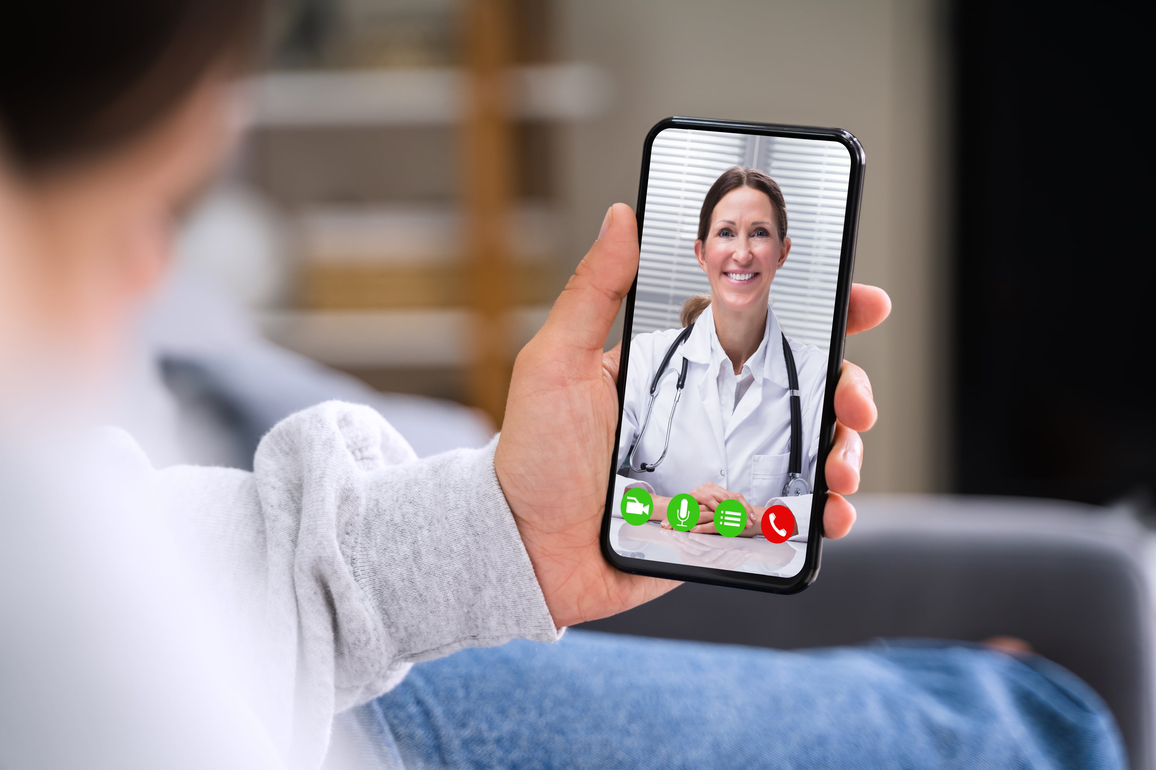 Person hält ein Smartphone in der Hand und führt ein Videotelefonat mit einer Ärztin in weißem Kittel und Stethoskop – typische Szene einer telemedizinischen Beratung.