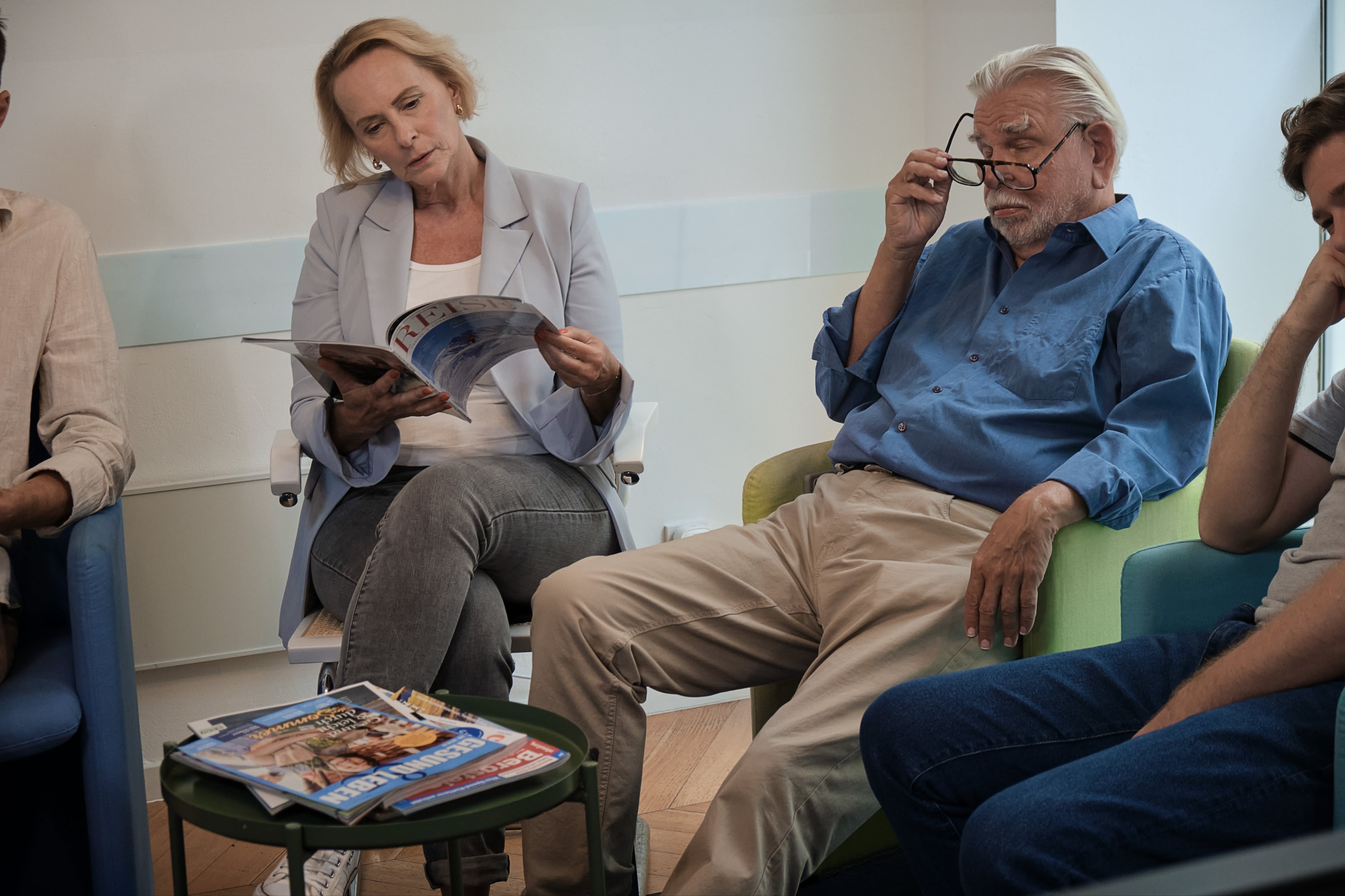 Mehrere Personen sitzen wartend in einem Wartezimmer. Eine Frau liest eine Zeitschrift, ein älterer Mann schaut über seine Brille, auf dem Tisch liegen weitere Magazine.