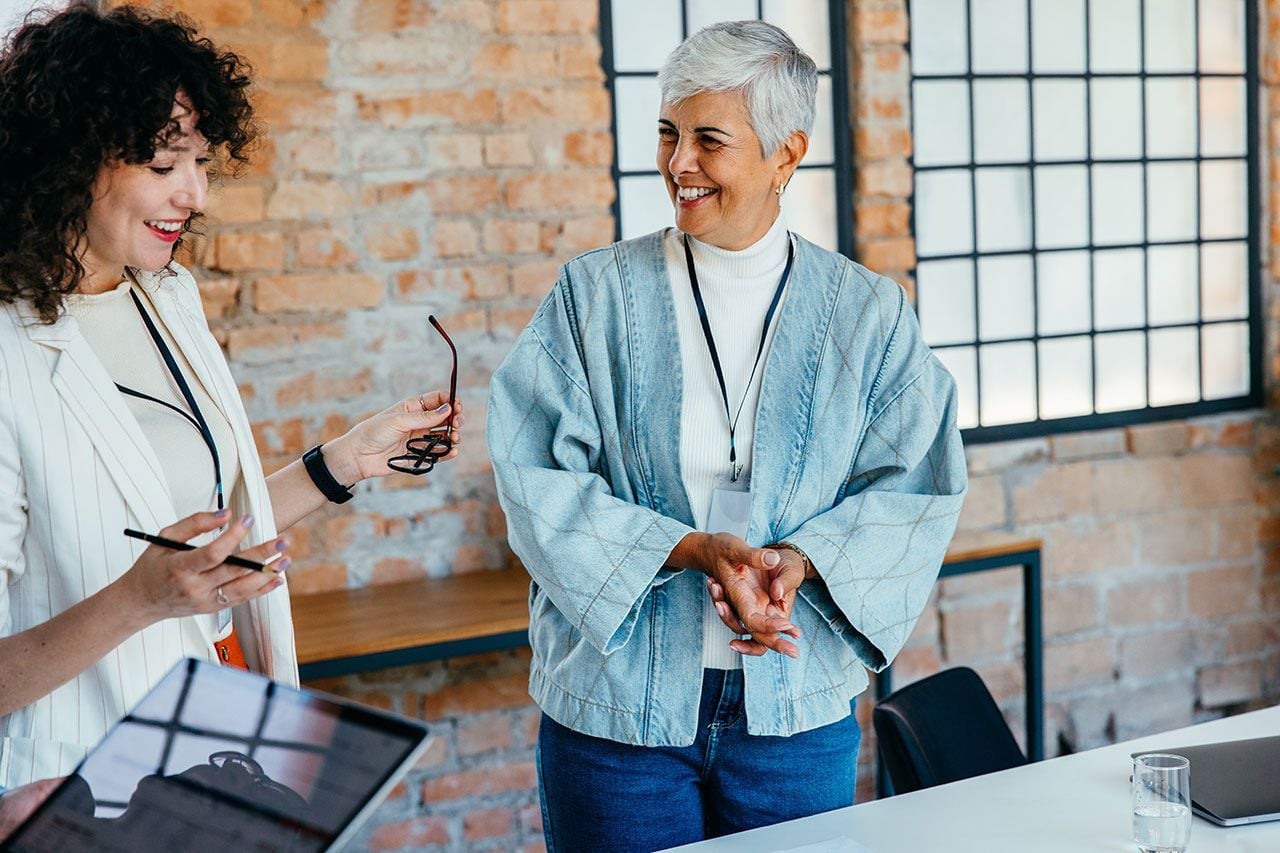 Zwei Frauen unterhalten sich lächelnd in einem modernen Büro mit Backsteinwand und großen Fenstern – eine entspannte Arbeitsatmosphäre.