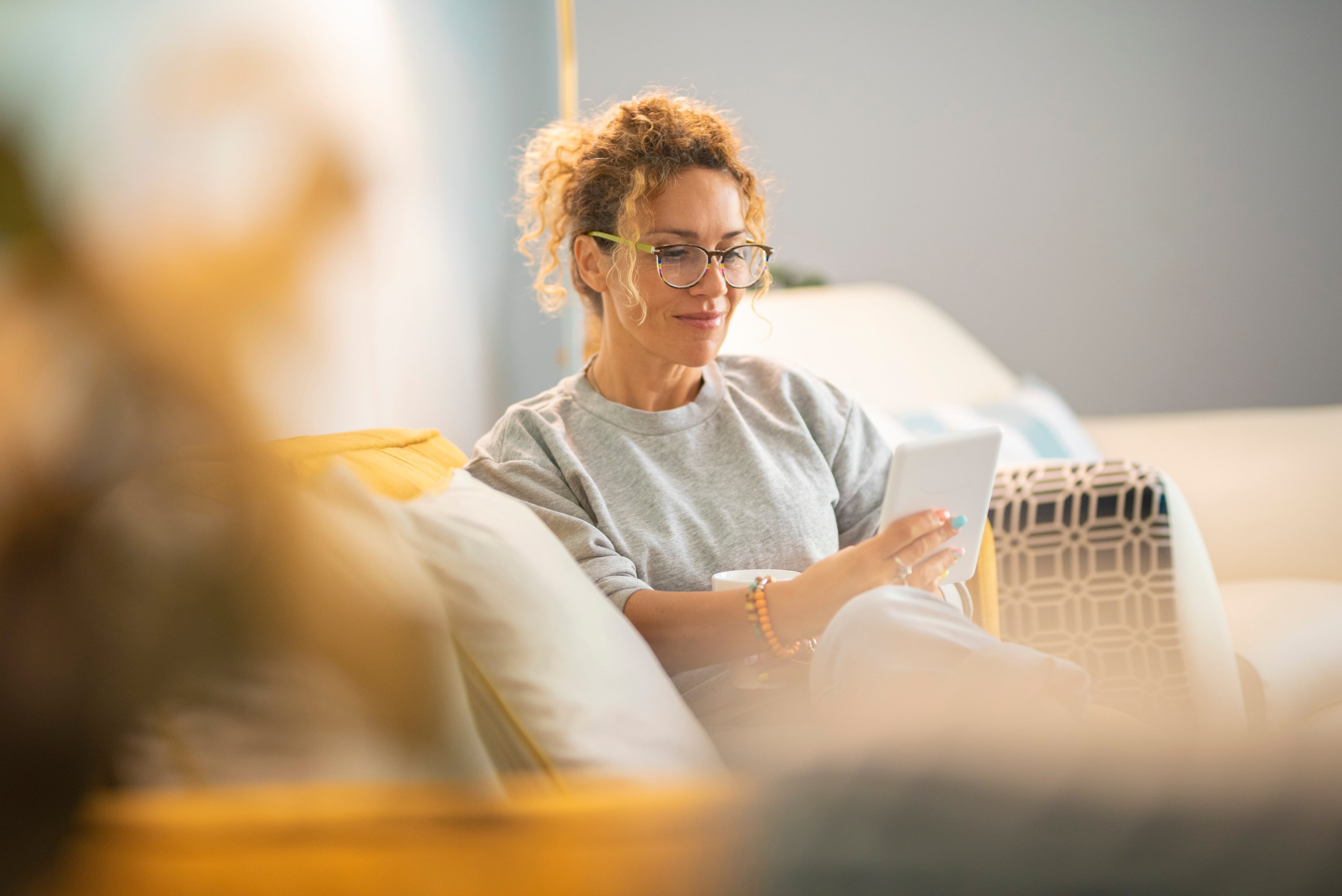 Frau mit lockigem Haar und Brille sitzt entspannt auf einem Sofa und liest auf einem Tablet. Die Atmosphäre ist ruhig und gemütlich, typisch für einen entspannten Moment zu Hause.