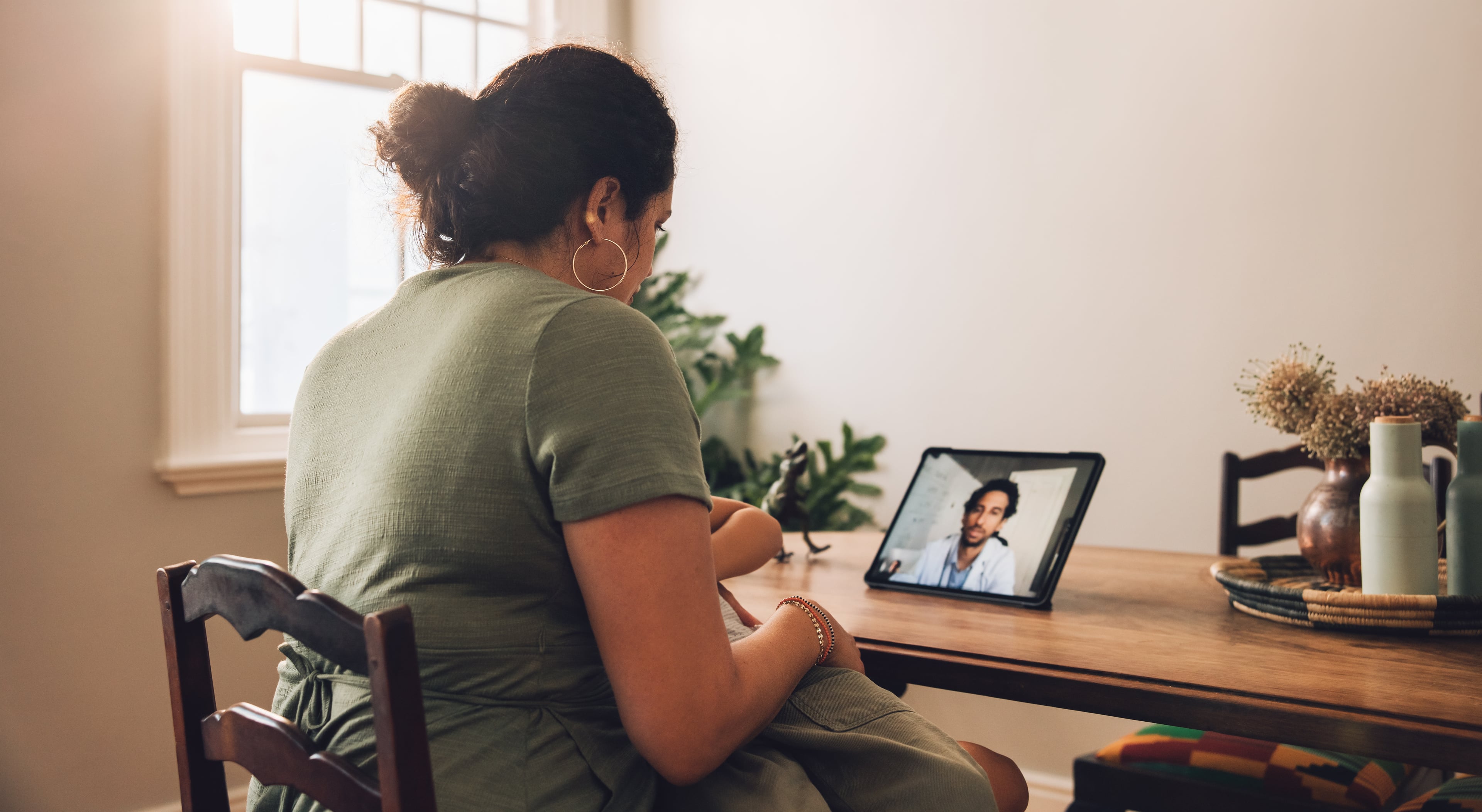Consultation GP Mavie Next Eine Frau sitzt mit einem Baby im Arm an einem Tisch und führt eine Videosprechstunde mit einem Arzt auf einem Tablet.