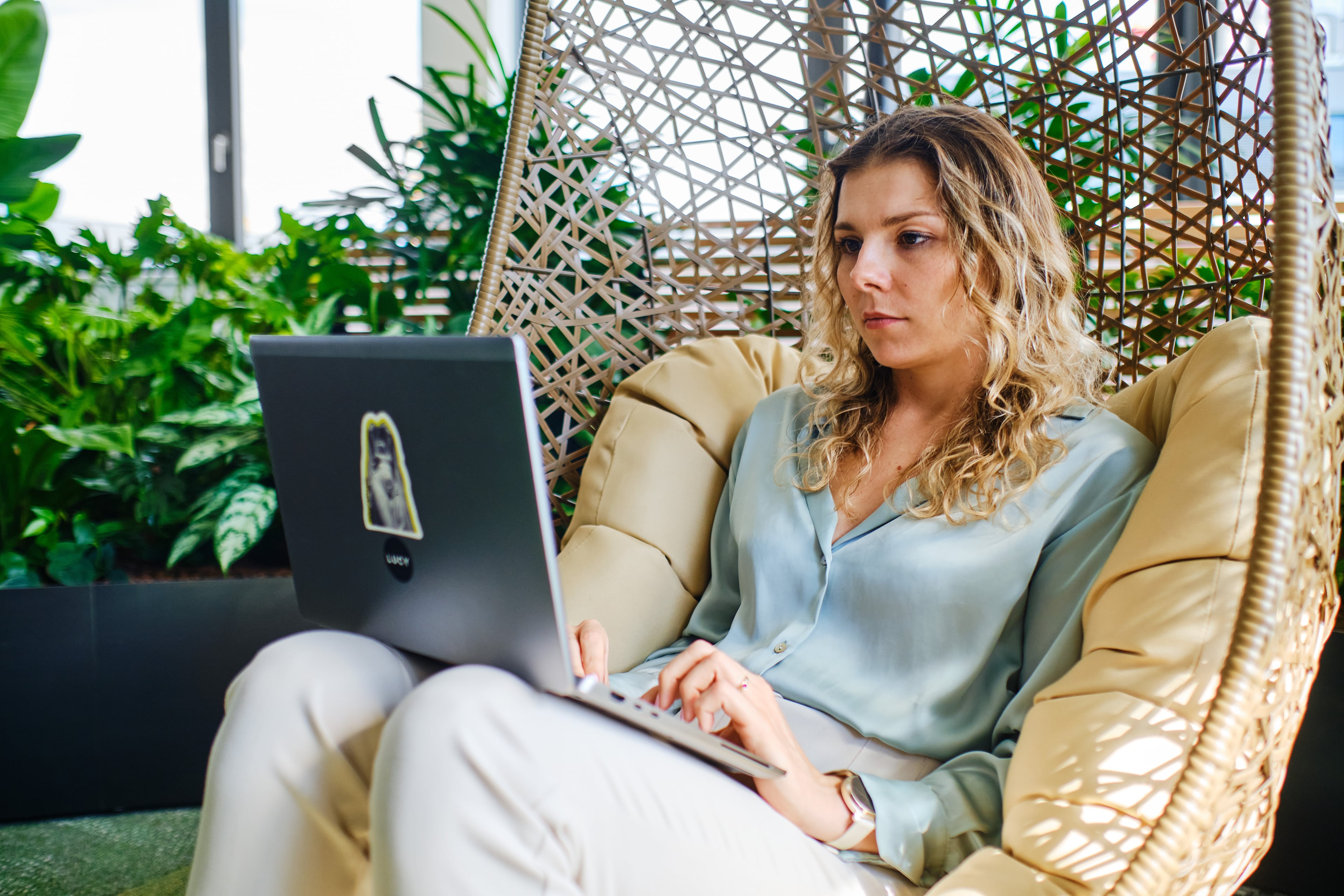Mental Health & Stress Studie Frau sitzt entspannt in einem Hängesessel und arbeitet konzentriert an einem Laptop. Um sie herum viele grüne Pflanzen – die Umgebung wirkt modern und naturnah.