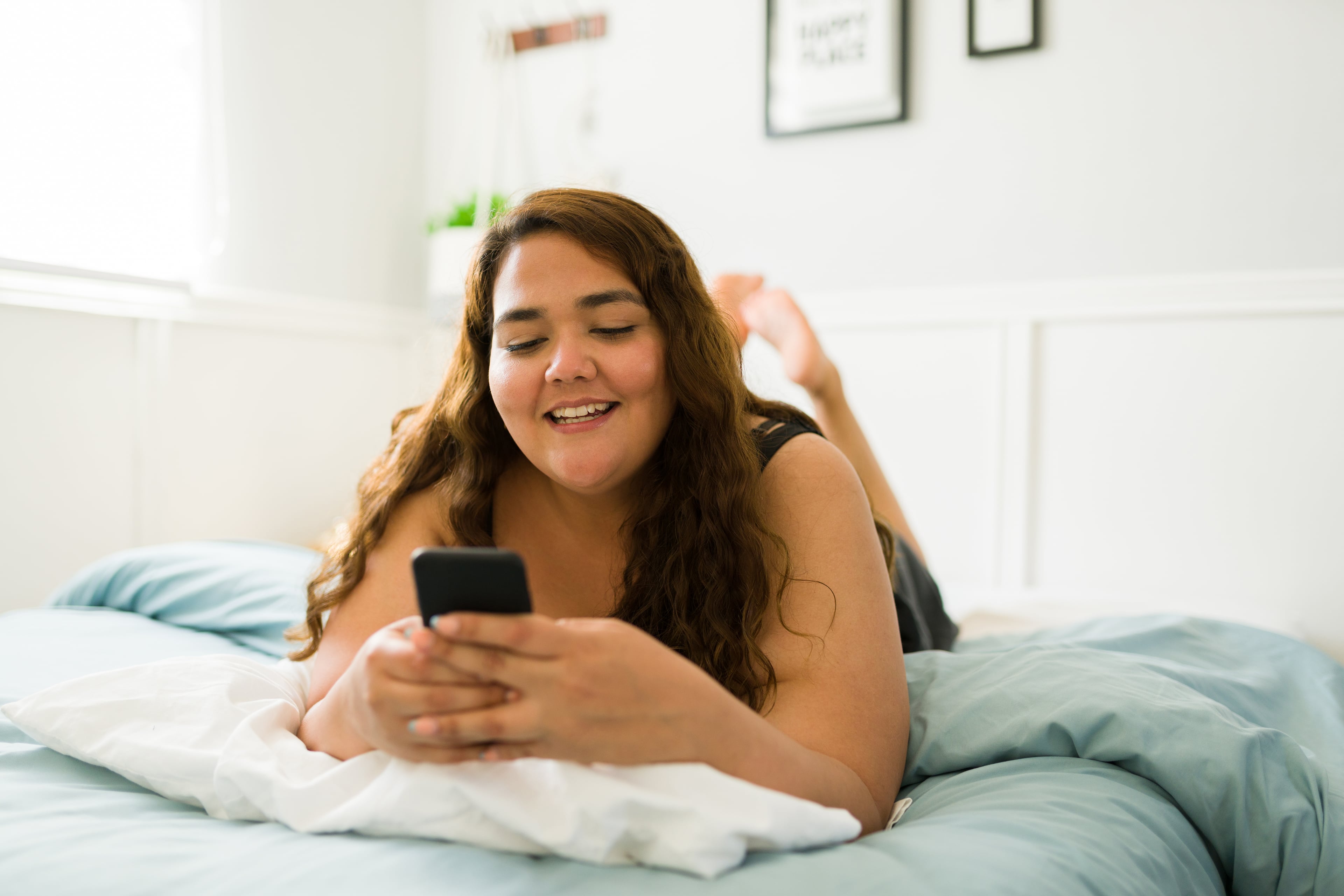 Woman lying on a bed and smiling while using her smartphone at home.
